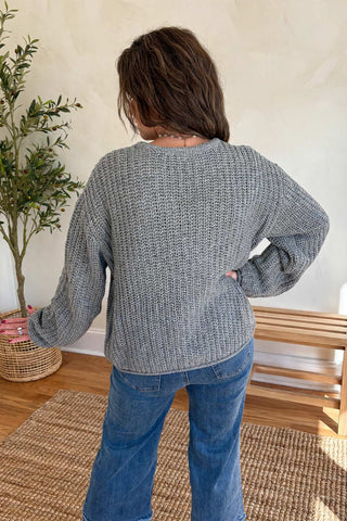 A person shown from the back wearing a chunky gray knit sweater and blue jeans in a bright indoor room with a plant, woven rug, and wooden bench.