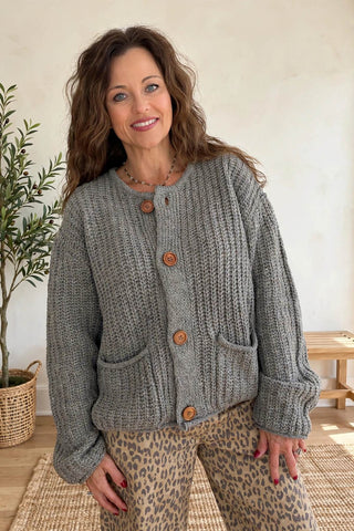 A woman wearing a chunky gray cable-knit cardigan with brown buttons and leopard-print pants, standing in a brightly lit room with a potted plant and wooden bench in the background.