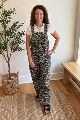 A woman modeling leopard-print overall jumpsuit with a white t-shirt underneath, standing in a bright room with a potted plant and wooden bench.