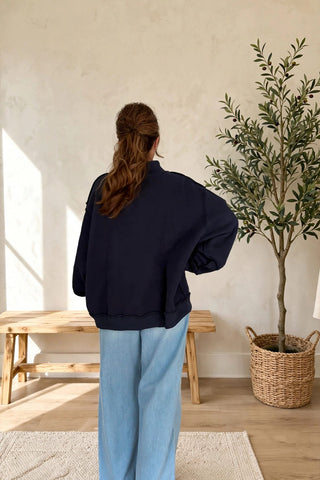 A woman viewed from behind wearing a dark navy blue oversized bomber jacket and light blue wide-leg jeans, standing in a bright room with a wooden bench, textured wall, and a potted olive tree.