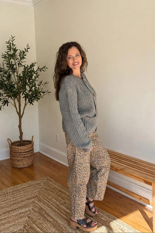 A woman stands in a bright room wearing a gray knit cardigan and beige leopard-print pants, with a potted tree and wooden bench in the background.