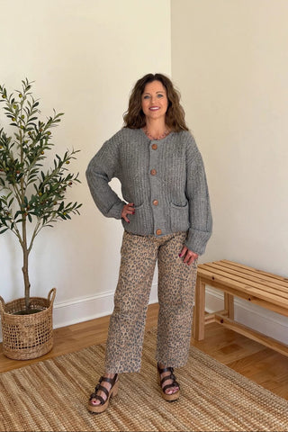 A woman modeling a gray knit button-front cardigan and beige patterned wide-leg pants in a bright room with a potted plant, woven rug, and wooden bench.