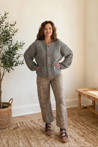 A woman modeling a two-piece knit loungewear set consisting of a gray button-up cardigan and beige patterned pants, standing in a brightly lit room with a potted plant and wooden bench.