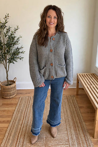 A woman modeling a chunky gray knit cardigan with brown buttons, paired with blue jeans and beige boots, standing on a woven rug in a bright room.