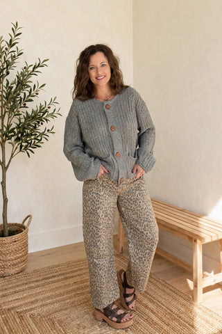 A woman stands in a bright room wearing a gray cable-knit button-front cardigan and beige patterned wide-leg pants, with sandals. A potted tree, woven rug, and wooden bench are visible in the background.