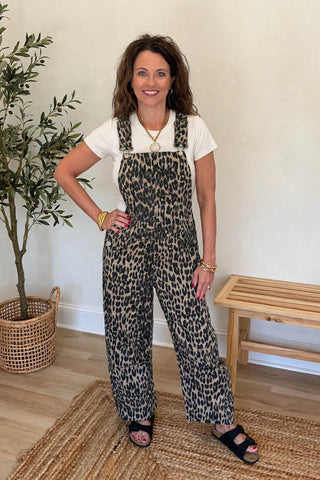 A woman modeling leopard-print overall-style jumpsuit over a white t-shirt, standing indoors on a woven rug next to a potted plant and wooden bench.
