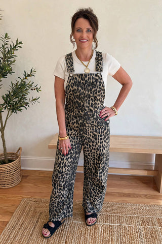 A woman modeling leopard-print overall sweatpants over a white t-shirt, standing in a brightly lit room with a wooden bench and potted plant in the background.