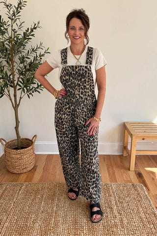 A woman modeling leopard-print overall jumpsuit over a white t-shirt, standing in a brightly lit room with a potted plant, wooden floor, and woven rug.