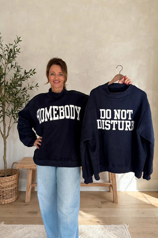 A woman stands indoors wearing a navy sweatshirt with white 'HOMEBODY' text, while holding up another navy sweatshirt on a hanger that reads 'DO NOT DISTURB'. She is also wearing light blue jeans, with a neutral wall, wooden stool, and potted plant in the background.