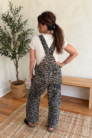 A woman stands with her back to the camera wearing leopard-print overall pants over a white t-shirt in a bright room with a potted plant and wooden bench.