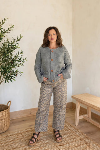 A woman stands in a bright room wearing a gray cable-knit cardigan with brown buttons and matching gray-and-beige patterned pants, paired with brown sandals.
