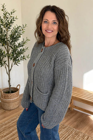 A woman wearing a chunky gray knit cardigan with brown buttons and blue jeans, standing in a brightly lit room with a potted plant and wooden bench in the background.