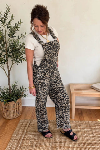 A woman modeling leopard-print overall jumpsuit with a white t-shirt and black slide sandals, standing on a woven rug in a room with a potted plant and a wooden bench.