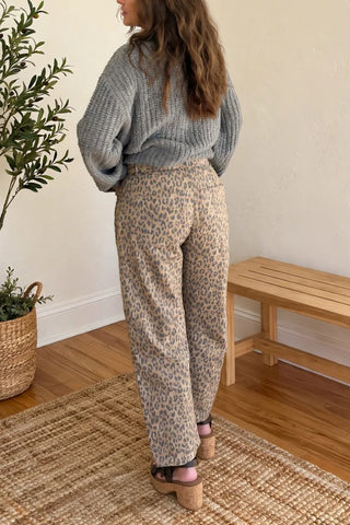 A woman stands with her back to the camera in a bright room, wearing a gray knit sweater and beige patterned wide-leg pants. A potted plant, a wooden bench, and a woven rug are visible in the room.