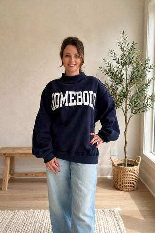 A woman wearing a navy blue sweatshirt with 'SOMEBODY' printed in white across the chest, paired with light blue jeans, standing in a bright room with a potted plant and wooden bench.