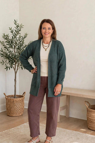 A woman modeling a dark green textured cardigan over a cream top and mauve-brown pants, standing in a bright, modern room with a potted olive tree and a wooden bench.