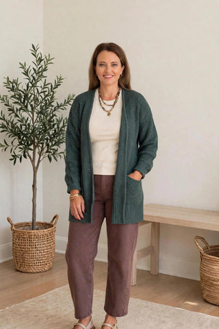 A woman modeling a dark green ribbed knit cardigan over a cream top and brown trousers, standing in a bright, modern room with a potted plant and wooden bench.