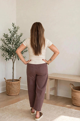A woman stands with her back to the camera wearing loose, wide-leg taupe-brown trousers and a cream short-sleeve top in a bright, minimalist room with a potted plant and a wooden bench.
