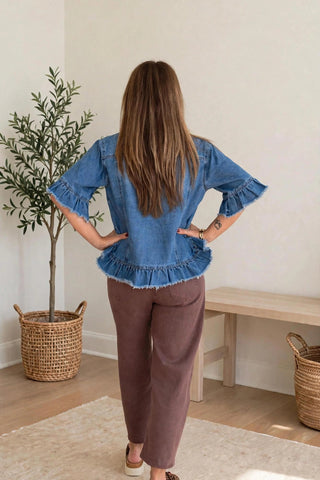 A woman viewed from behind wearing a blue denim top with ruffled sleeves and hem, paired with brown pants, standing in a bright room with a potted plant and wooden bench.