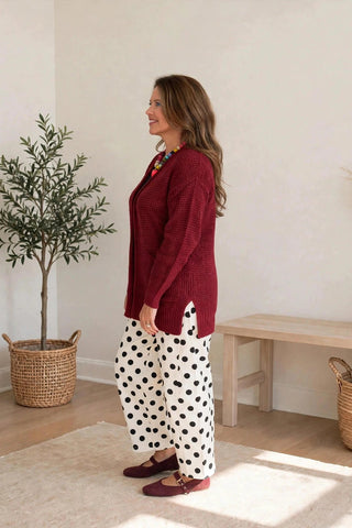A woman stands in a bright room wearing a burgundy knit cardigan, white pants with black polka dots, and burgundy flats. A potted tree, a wooden bench, and woven baskets are visible in the background.
