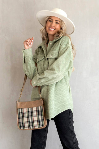 Woman wearing a green coat and white hat, holding a plaid bag against a plain background