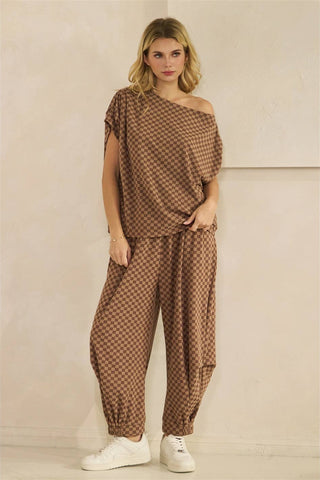 A woman modeling a brown and beige checkered two-piece outfit consisting of an asymmetrical draped top and matching loose pants, paired with white sneakers against a neutral beige wall.