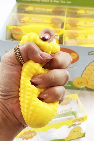 A hand squeezing a yellow textured squishy toy shaped like a corn cob, with a matching product box visible in the background.