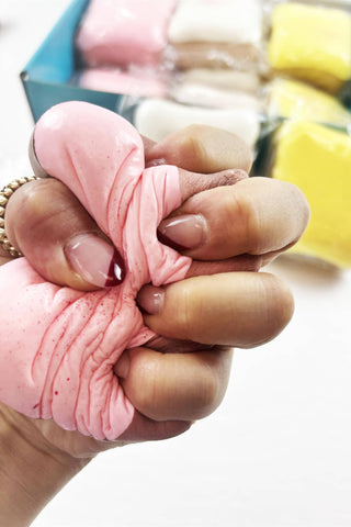 A close-up of a hand squeezing a pink squishy toy, with a box of colorful squishies in the blurred background.