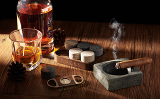 Cigar and cigar accessories on a wooden table with a bottle of alcohol and glass.