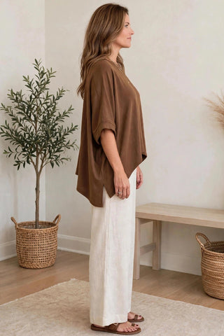 A woman stands in profile wearing a loose brown short-sleeve top and off-white wide-leg pants in a bright indoor room with a plant, bench, and woven baskets.