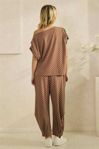 A woman viewed from behind wearing a brown and beige checkered two-piece lounge set with an off-the-shoulder top and matching loose pants, paired with white sneakers in a softly lit indoor setting.