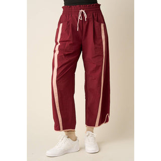 A person wearing loose burgundy cropped pants with light beige side stripes, a white drawstring waist, and white sneakers, shown against a plain beige studio background.