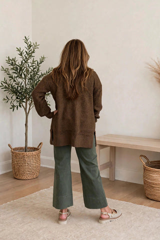 A woman stands with her back to the camera in a bright, minimalist room, wearing a brown textured knit cardigan, olive green wide-leg pants, and pink sandals.