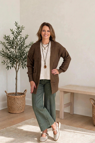 A woman modeling a brown plaid cardigan over a cream top and olive green wide-leg pants, standing in a bright, minimalist room with a potted tree and a wooden bench.