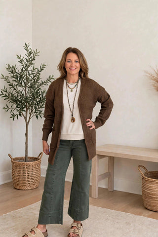A woman modeling a brown knit cardigan over a cream top and green wide-leg pants, standing in a bright room with a potted plant, wooden bench, and woven baskets.