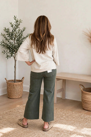 A woman stands with her back to the camera in a bright, minimalist room, wearing a cream-colored long-sleeve top and olive green wide-leg pants.