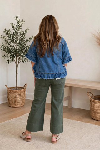 A woman viewed from behind wearing a blue denim jacket with ruffled sleeves and hem, paired with olive green wide-leg pants and pink sandals, standing in a bright room with a potted plant and wooden bench.