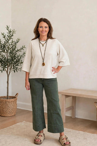 A woman modeling a cream-colored short-sleeve top and dark green cropped pants, standing in a minimalist room with a potted plant, wooden bench, and woven baskets.
