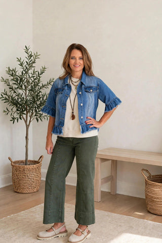 A woman modeling a blue denim jacket with ruffled sleeves, worn over a cream t-shirt and paired with olive green wide-leg pants and beige shoes, standing in a bright, modern room with a potted plant and wooden bench.