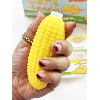 A hand with manicured nails holds a yellow corn-shaped squishy toy, with a cartoon corn box blurred in the background.
