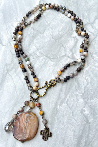 A beaded necklace with a large oval agate pendant, featuring multi-strand beads in earthy tones of brown, gray, white, and yellow, with a brass clasp and hanging charms, displayed on a textured white fabric background.
