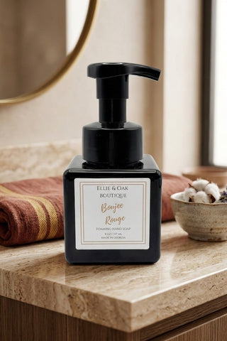 A black pump bottle of Ellie & Oak Boutique Boujee Rouge hand soap standing on a beige stone countertop in a bathroom setting, with a folded rust-colored towel, a small bowl of cotton buds, and a mirror in the background.