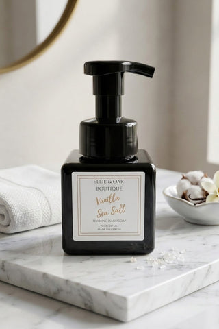 A black pump bottle of Ellie & Oak Boutique Vanilla Sea Salt hand soap resting on a white marble tray, styled with a folded white towel and a small bowl of cotton buds in a bright bathroom setting.