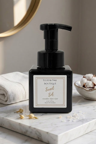 A black pump bottle of Ellie & Oak Boutique Sweet Sol hand soap sits on a white marble tray, styled with a folded white towel, gold decorative accents, and a small bowl of cotton bolls in a bright bathroom setting.