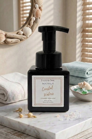 A black pump bottle of Ellie & Oak Boutique Coastal Waters coconut soap sits on a marble countertop in a bathroom setting with folded towels, a decorative mirror, and seashell accents.