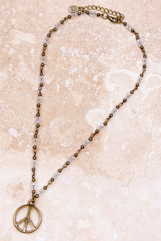 Gold beaded peace sign necklace with clear crystal beads and bronze pendant on blush marble surface