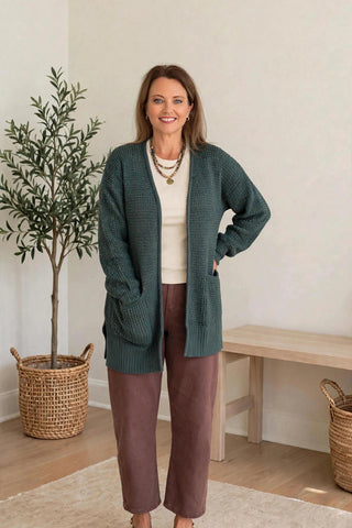 A woman modeling a dark teal green waffle-knit cardigan over a cream top and brown pants, standing in a bright room with a potted plant and a wooden bench.