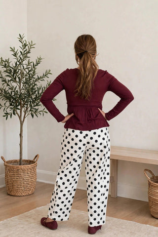 A woman stands with her back to the camera in a bright room, wearing a burgundy long-sleeve peplum top and white pants with black polka dots.