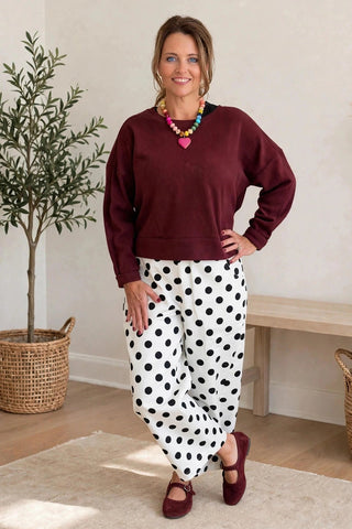A woman modeling a two-piece outfit consisting of a maroon long-sleeve cropped sweatshirt and white pants with black polka dots, standing in a brightly lit room with a potted olive tree and a wooden bench.