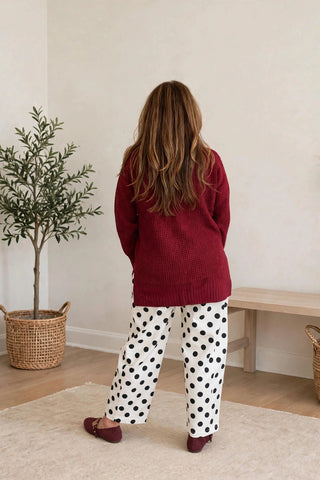 A woman stands with her back to the camera in a cozy room, wearing a ribbed burgundy sweater and white pajama pants with black polka dots. She is wearing burgundy flats and standing on a light beige rug next to a potted olive tree and a wooden bench.